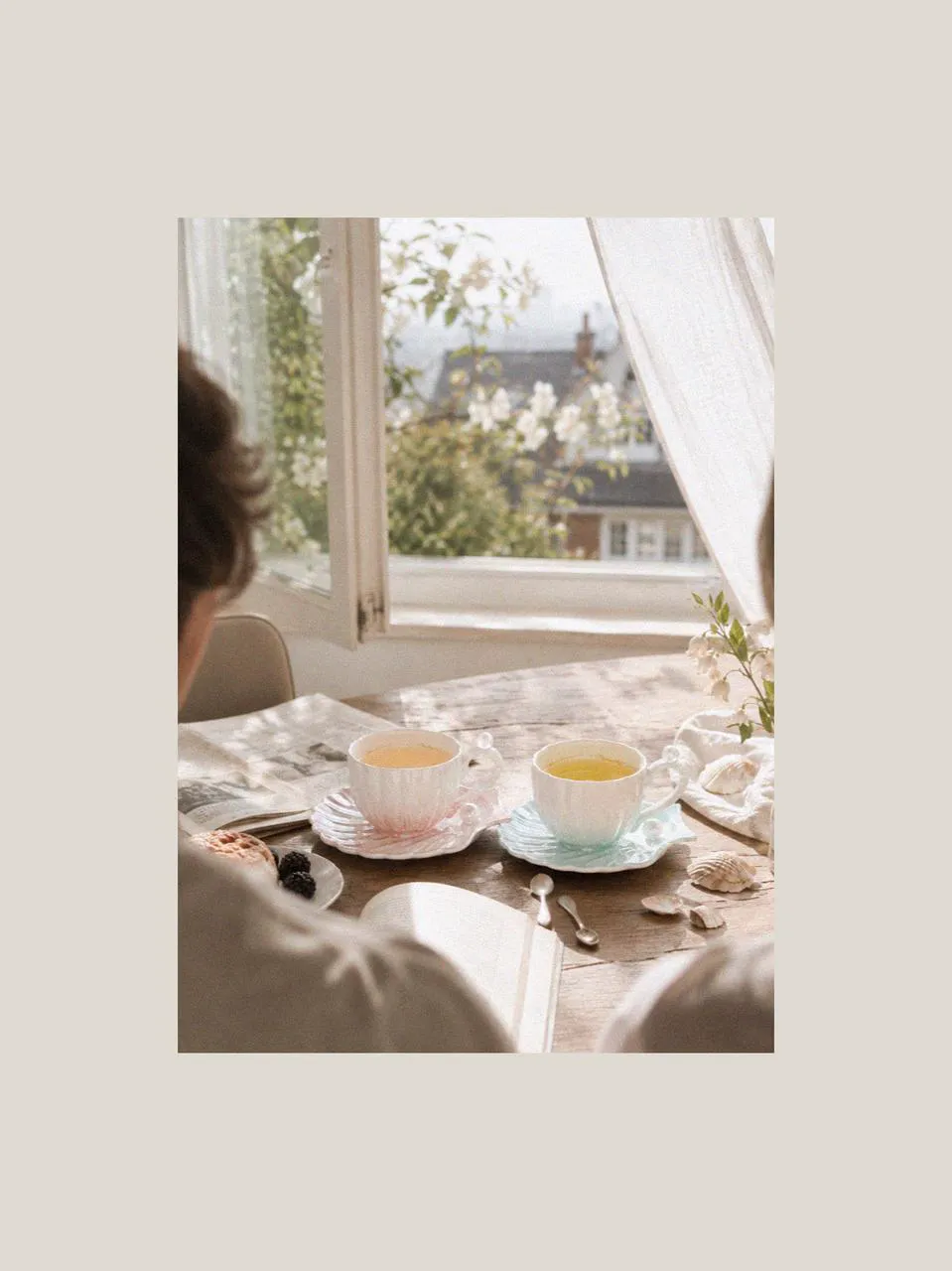 A ceramic mug set with pastel shell-inspired saucers on a sunlit table beside open windows, books, and delicate beach shells, creating a calm coastal atmosphere for shared moments in London during summer.